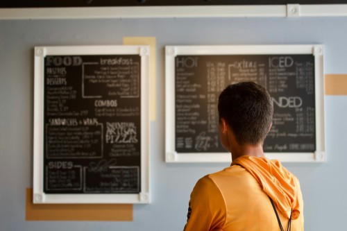 A boy in a yellow top an brown hair faces a chalkboard menu on the wall.